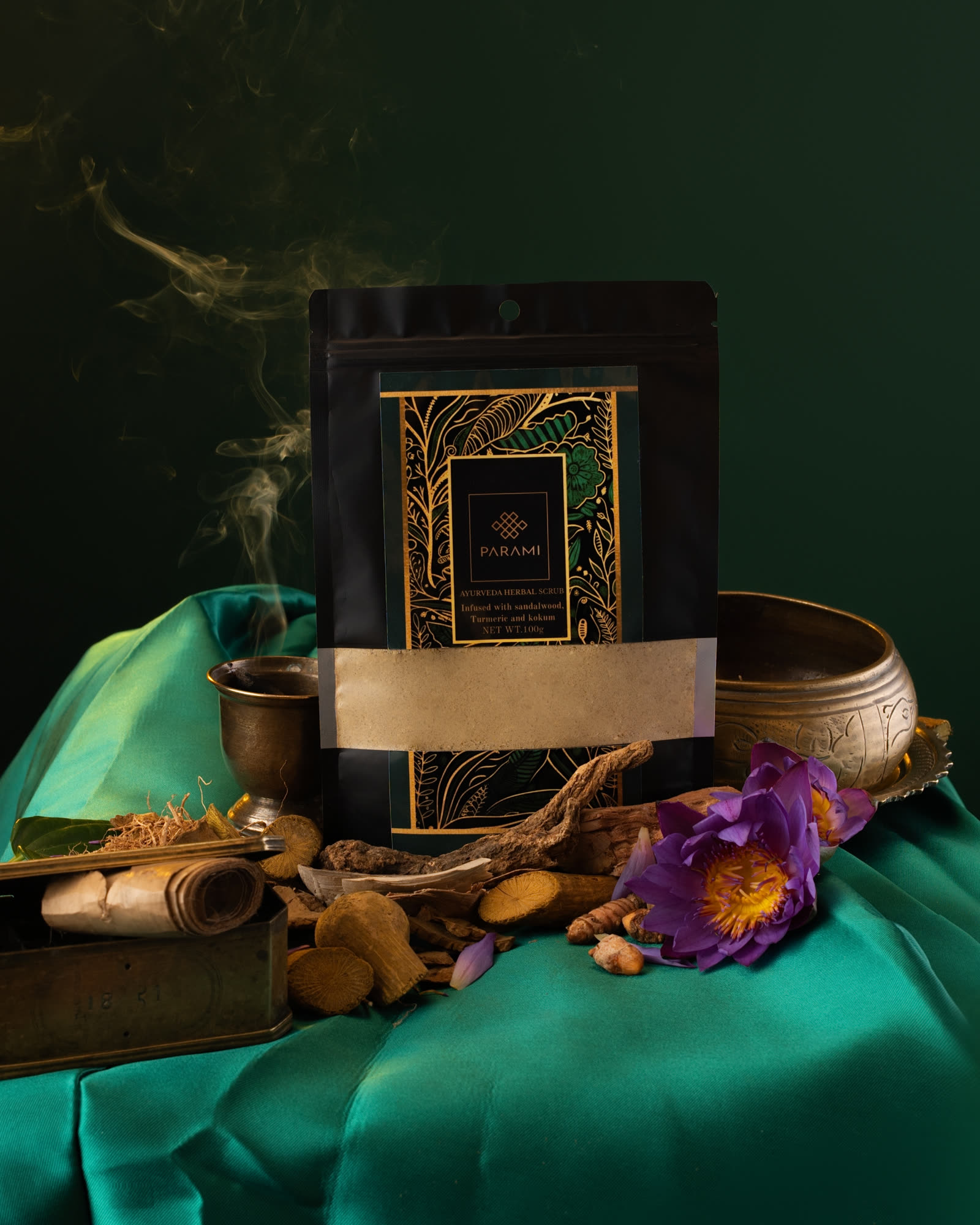 The Parami Ayurveda Herbal Scrub pouch on green silk with roots, a brass bowl, a blue water lily and a thin rise of incense smoke