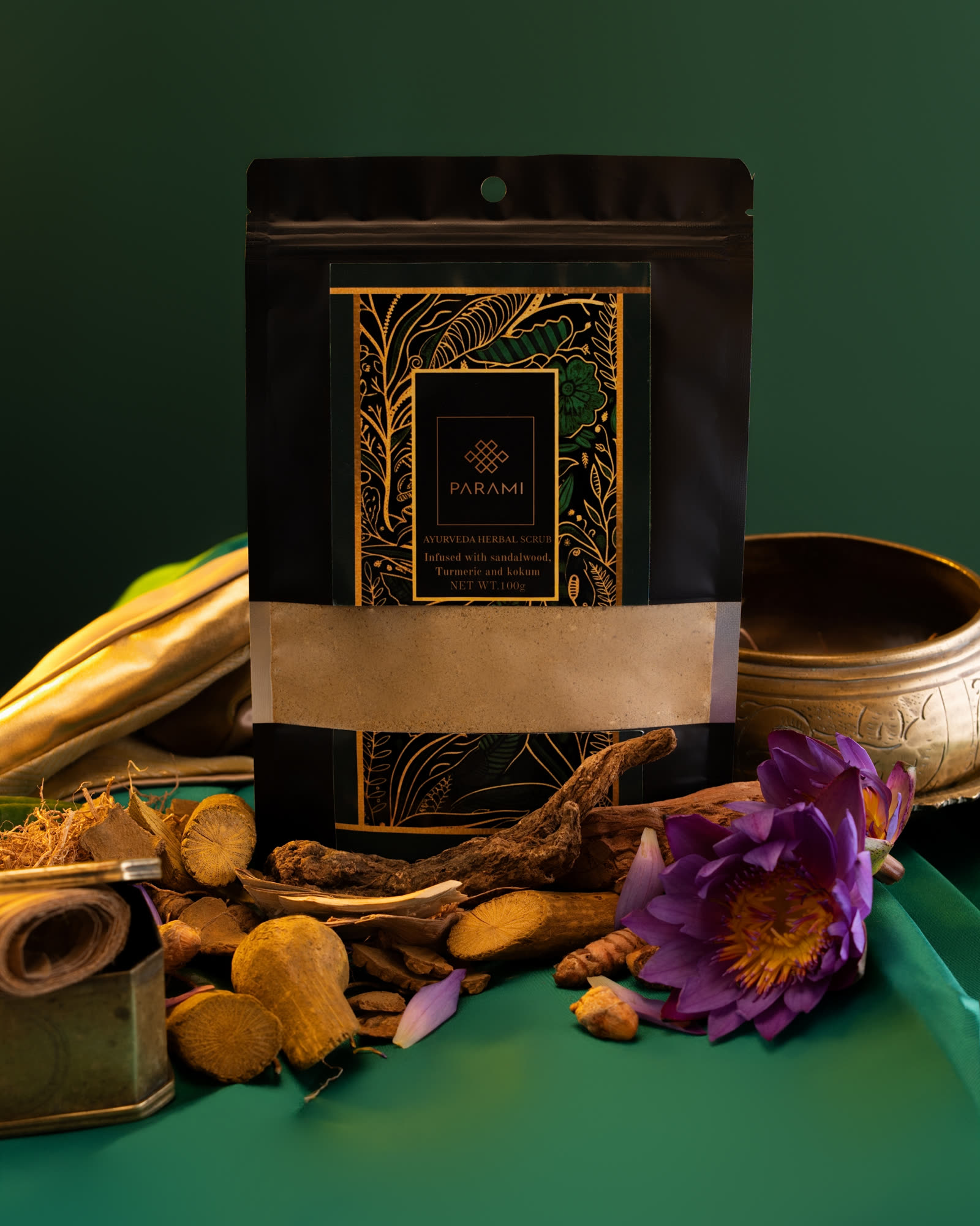 The Parami Ayurveda Herbal Scrub pouch, black with gold botanical line work, among sandalwood, turmeric root, a singing bowl and a blue water lily on green silk